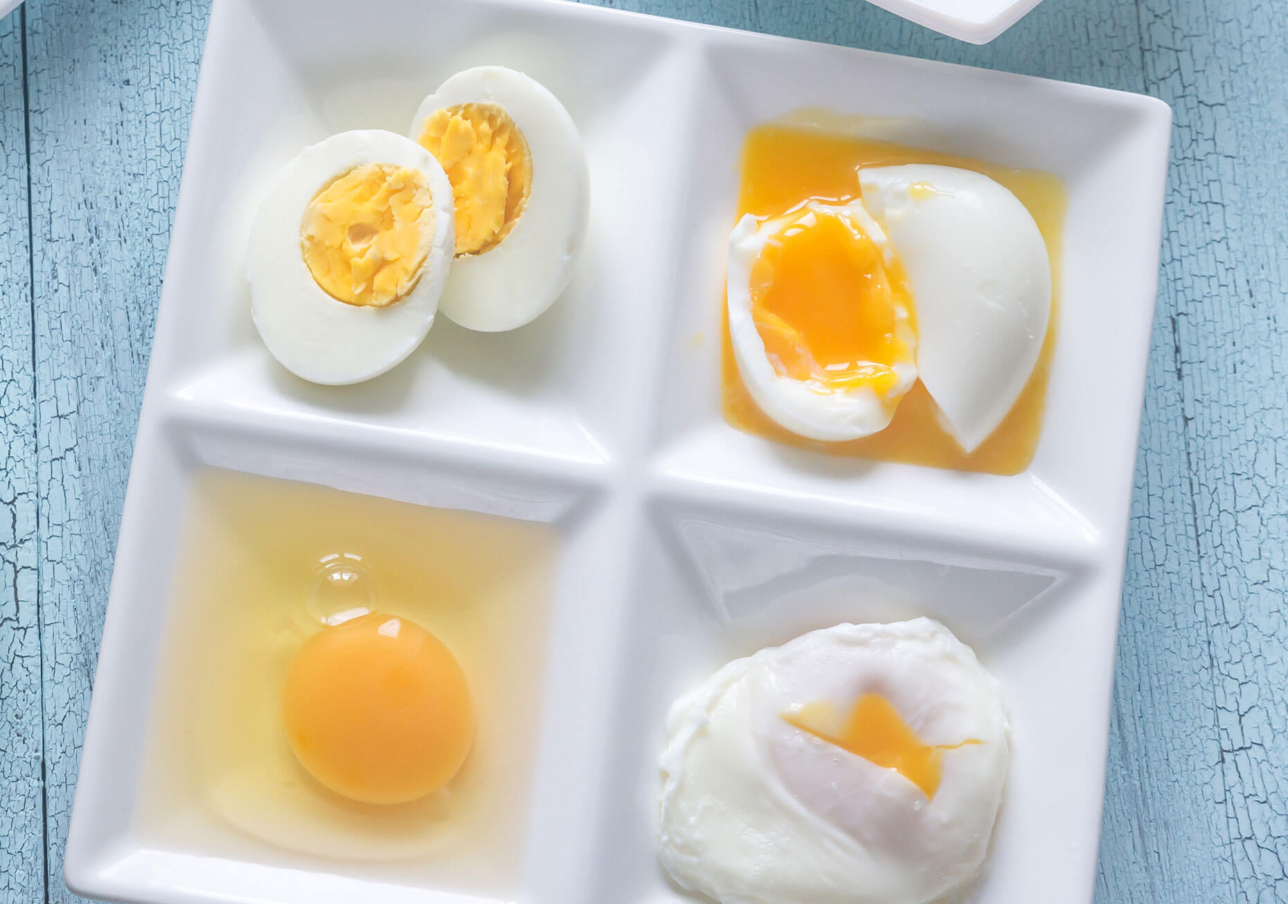 Four eggs in each corner of a square plate; clockwise from upper left: hard-boiled, soft-boiled, poached, and raw