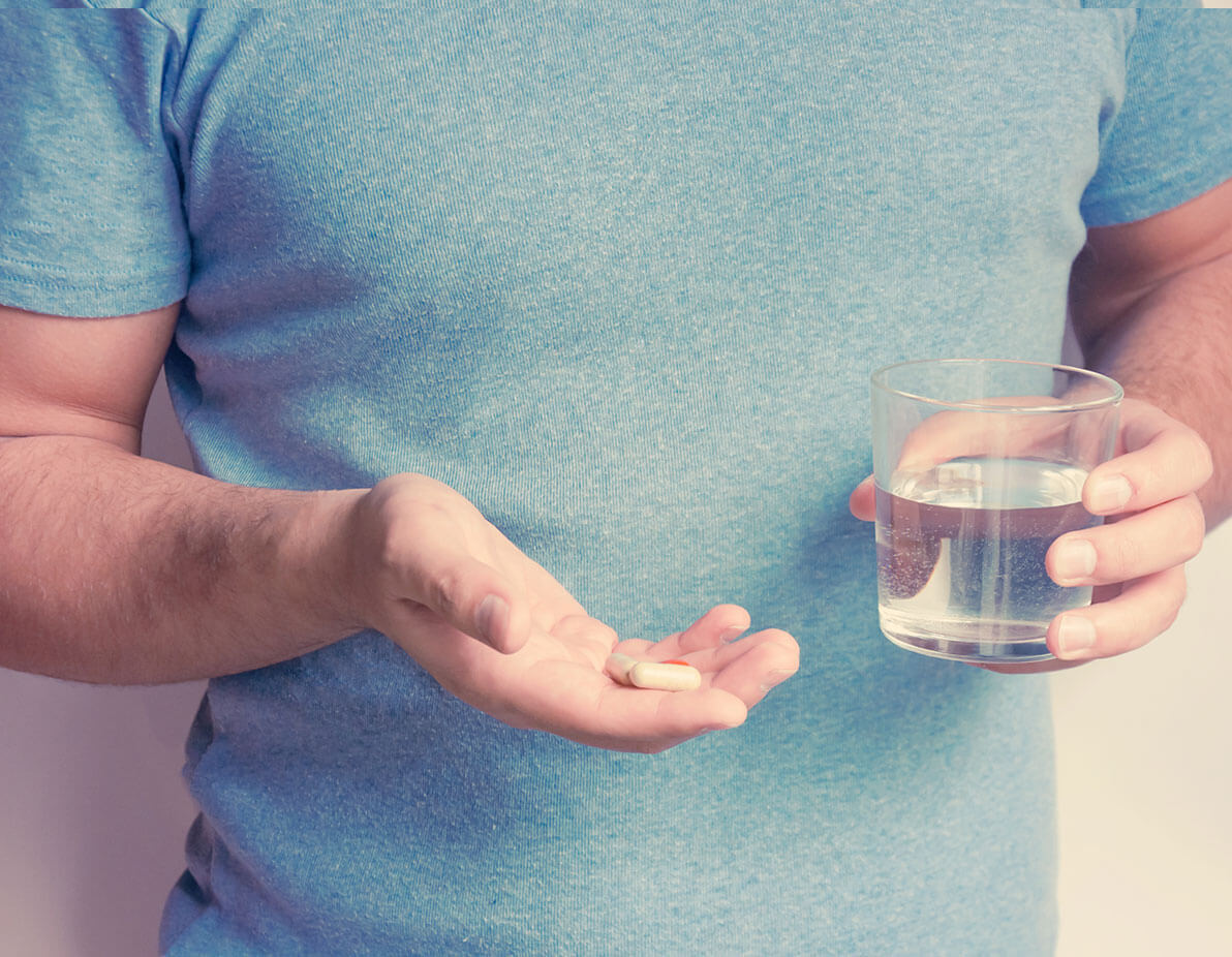 A torso shot of a man in a blue t-shirt, holding a glass of water and two capsules