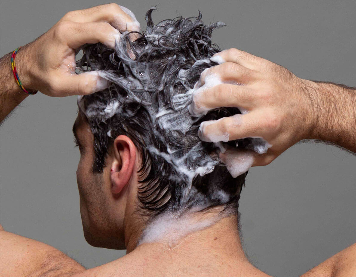 A man facing backwards, with his hands in his hair as he shampoos it