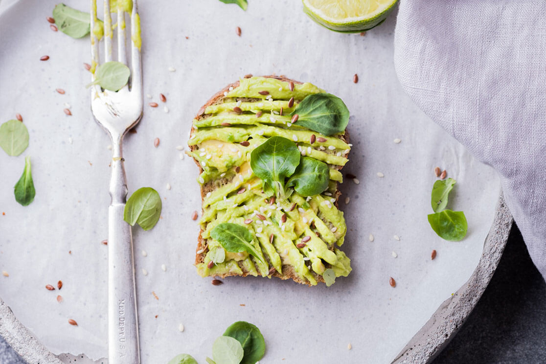 Smashed avocado on toast in the center of a gray plate, with a fork to the left, halved lime above, and baby spinach scattered around it