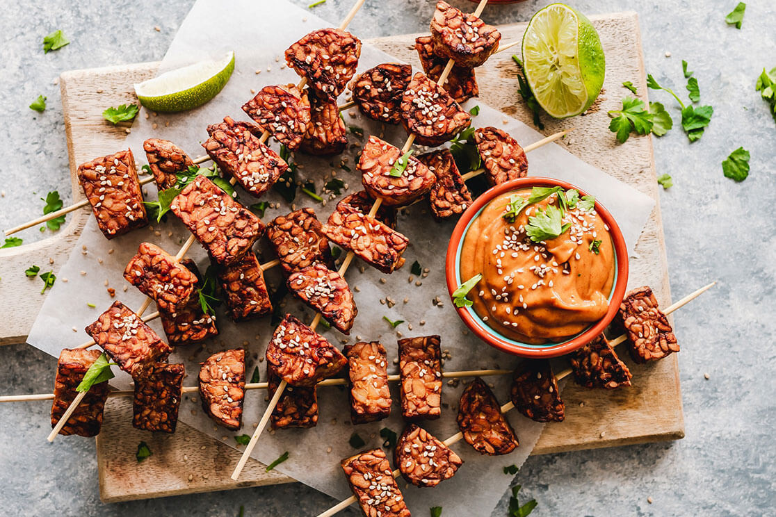 Six tempeh and peanut skewers on a wooden chopping board, alongside slices of lime and a container of creamy peanut satay dip